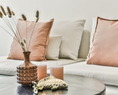 Setting mit apricotfarbenen Kissen auf kreidefarbenem Sofa, runder Tisch mit Vase mit Gräsern, 2 farblich passenden Kerzen